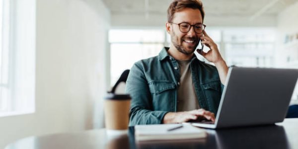 Software designer discussing a new project with his client on the phone. Creative business man working on a laptop in an open plan office.